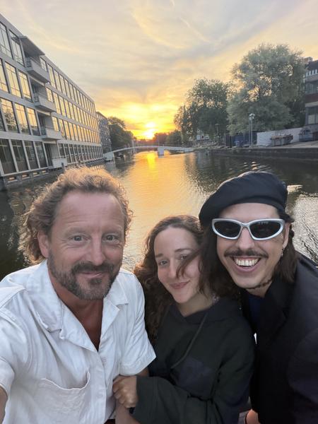 Three people are captured in a joyful selfie, with two smiling broadly and one with a gentle smile, all bathed in the warm glow of a sunset